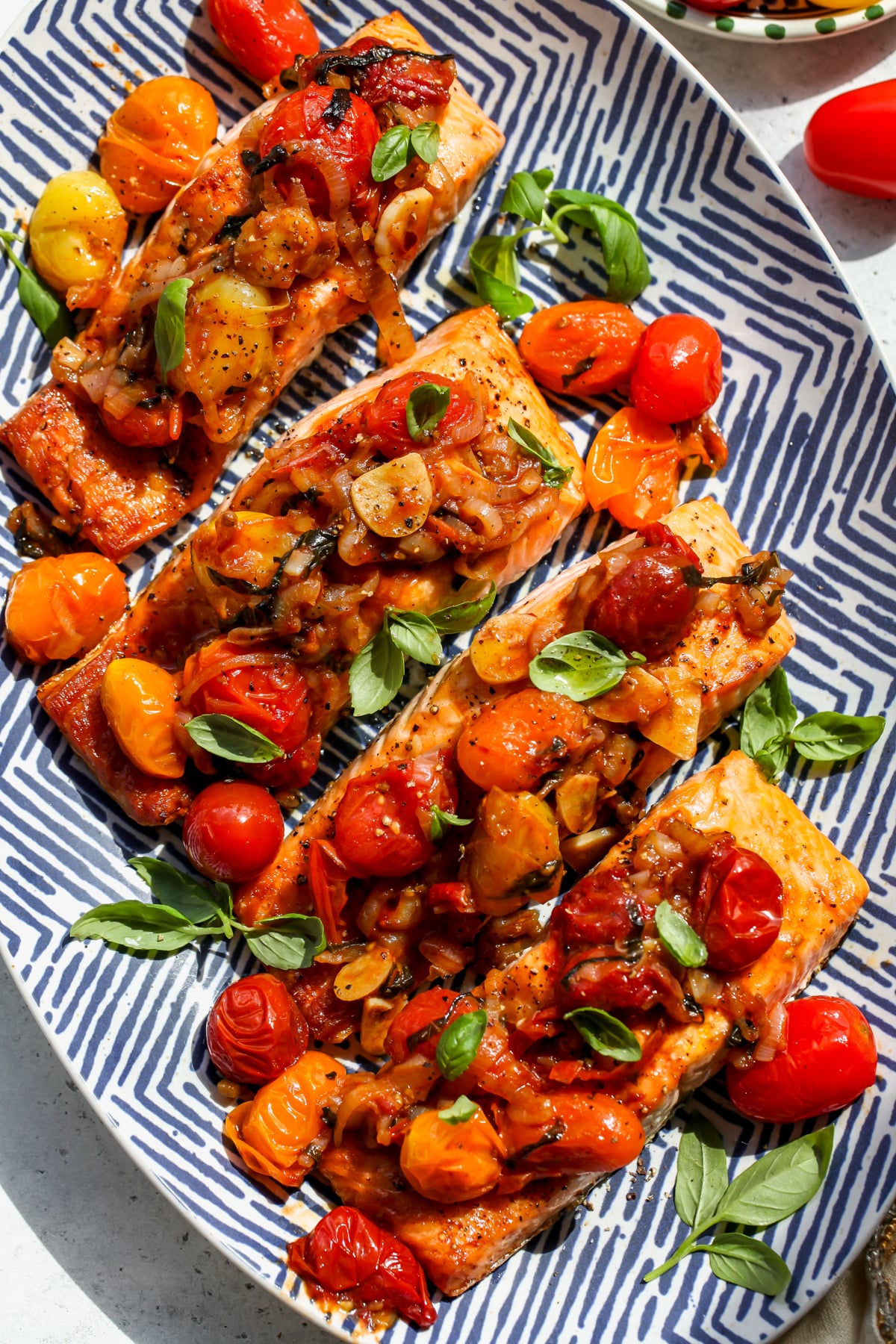 Four broiled salmon fillets topped with balsamic tomatoes on a blue and white platter.