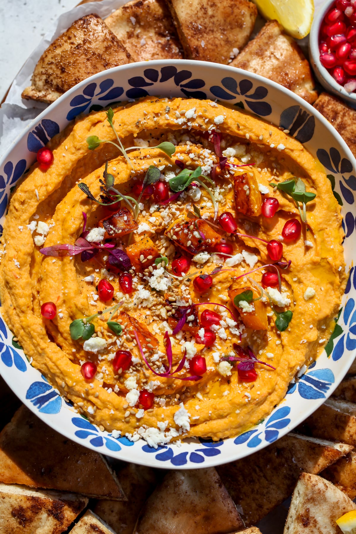 Butternut squash hummus topped with pomegranate, feta, microgreens, and sesame seeds in a blue and white bowl.