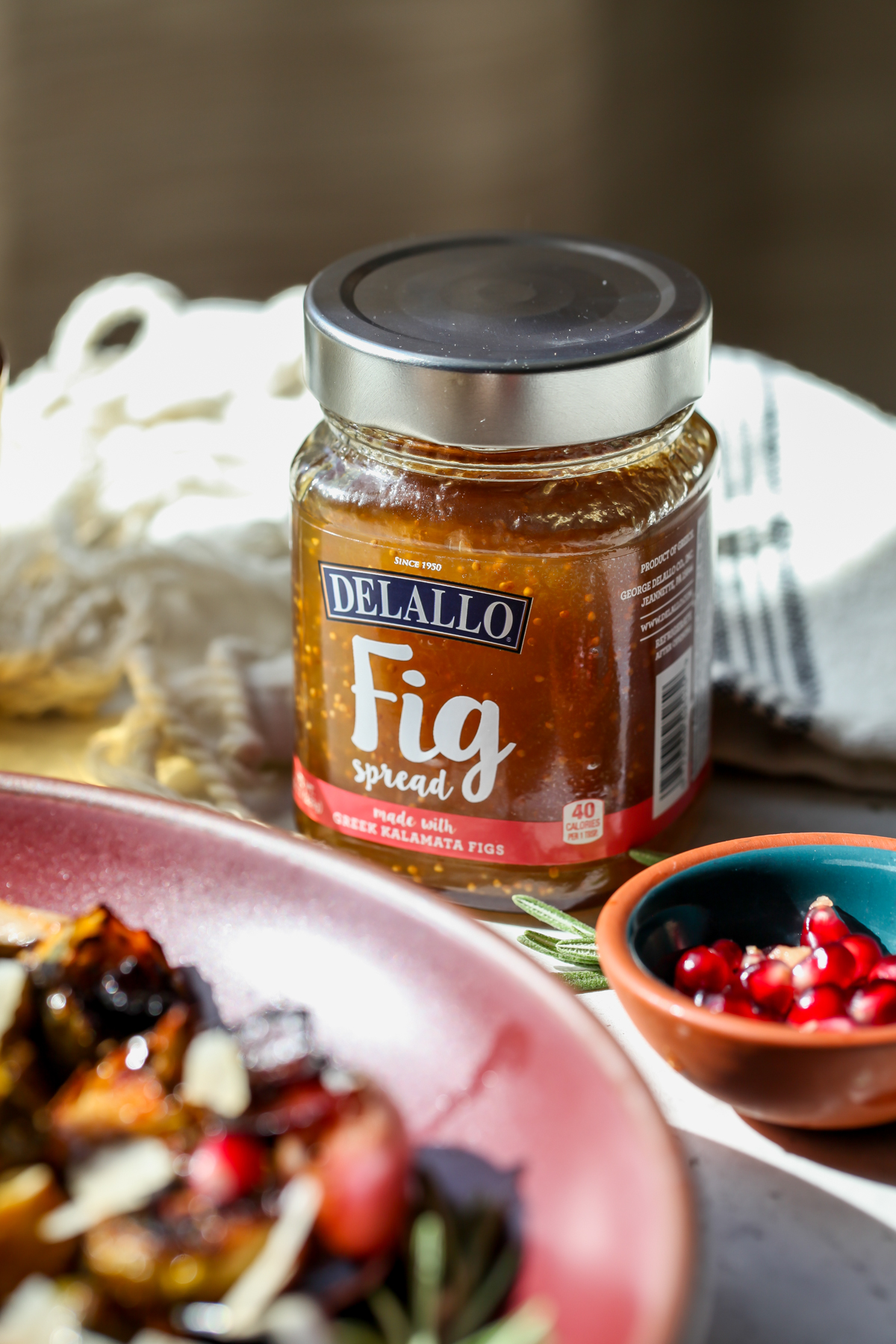 A jar of DeLallo Fig spread on a surface with a bowl of pomegranate arils on the right side.