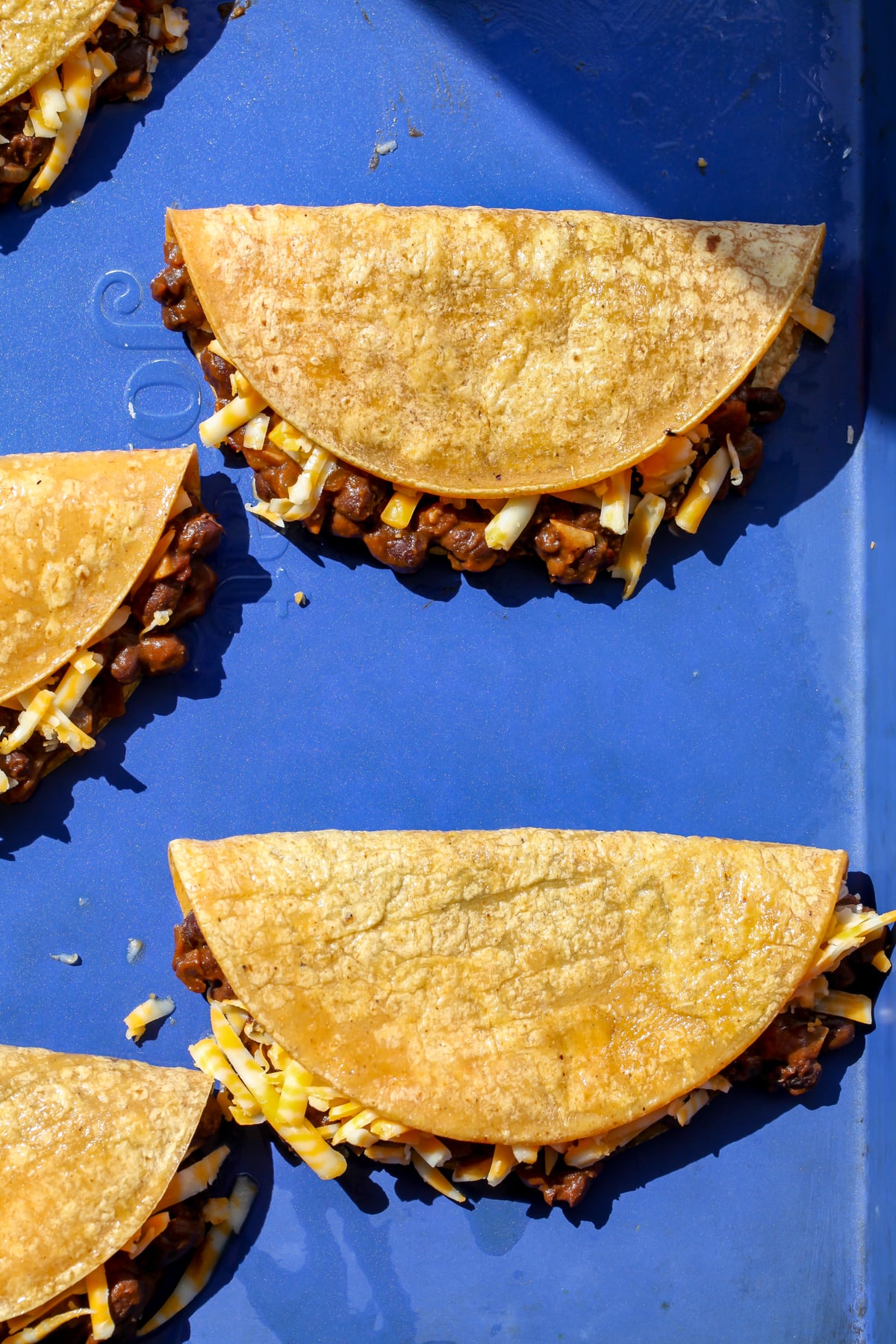 Filling and cheese being added to corn tortillas arranged on a large rimmed baking sheet.
