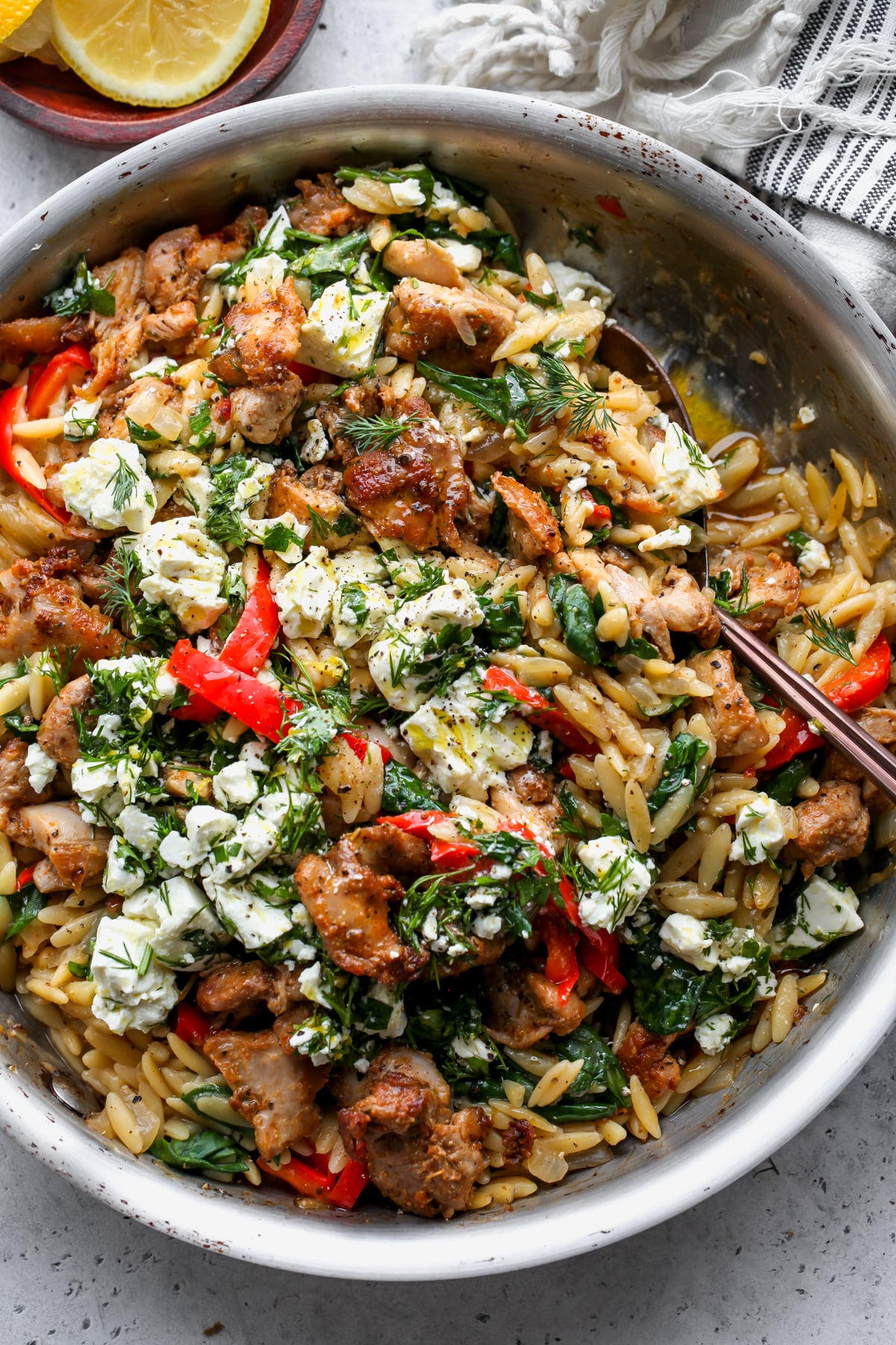 Chicken and orzo topped with marinated feta in a skillet. Serving spoon resting on the right side.