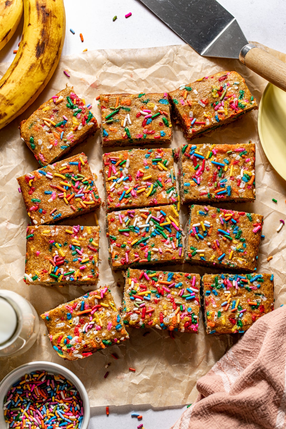 Sliced banana bread bars arranged on a work surface surrounded by ripe bananas and a bowl of sprinkles.