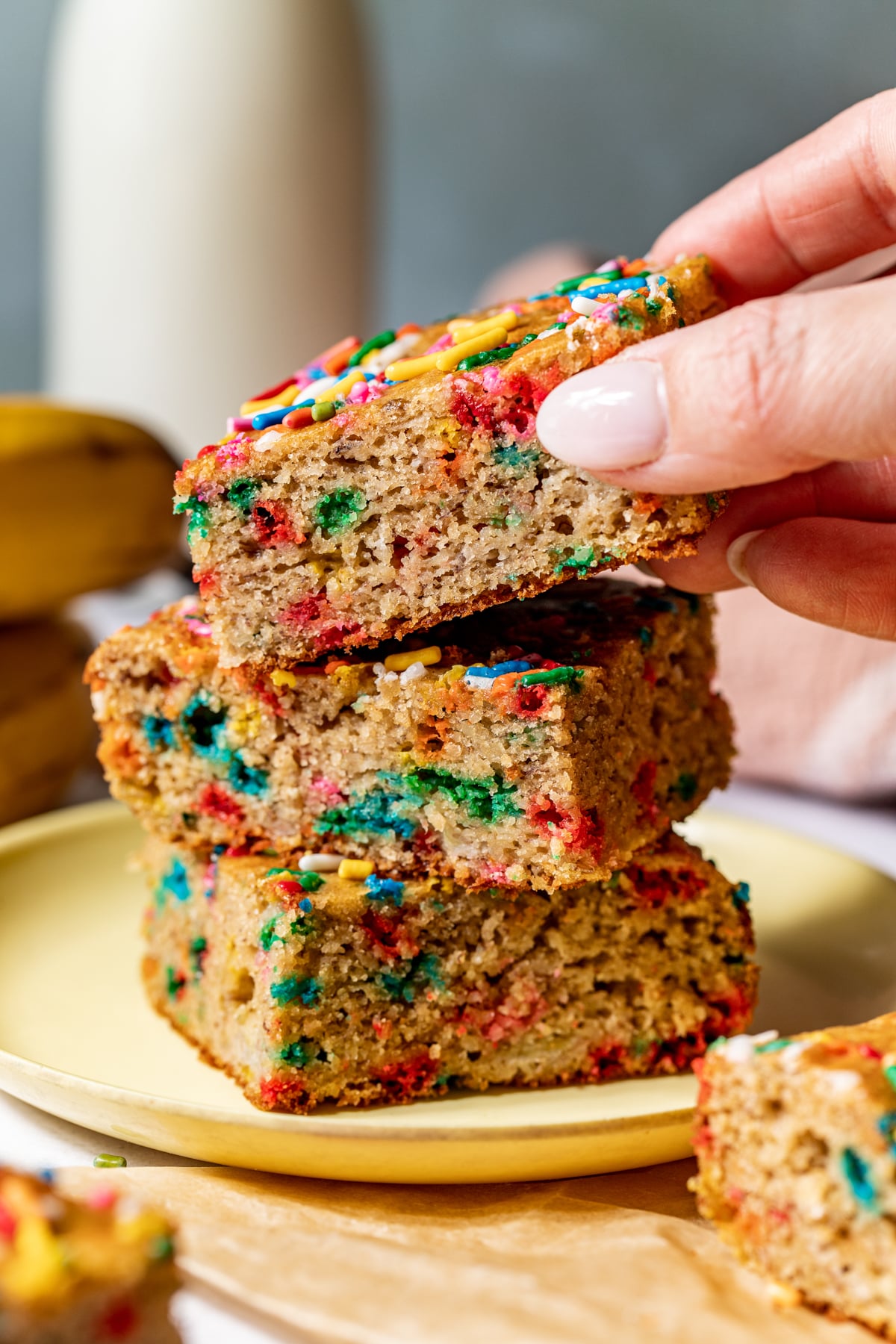 Healthy funfetti bars stacked on top of one another on a small yellow plate. A hand reaching in to take the bar on top.