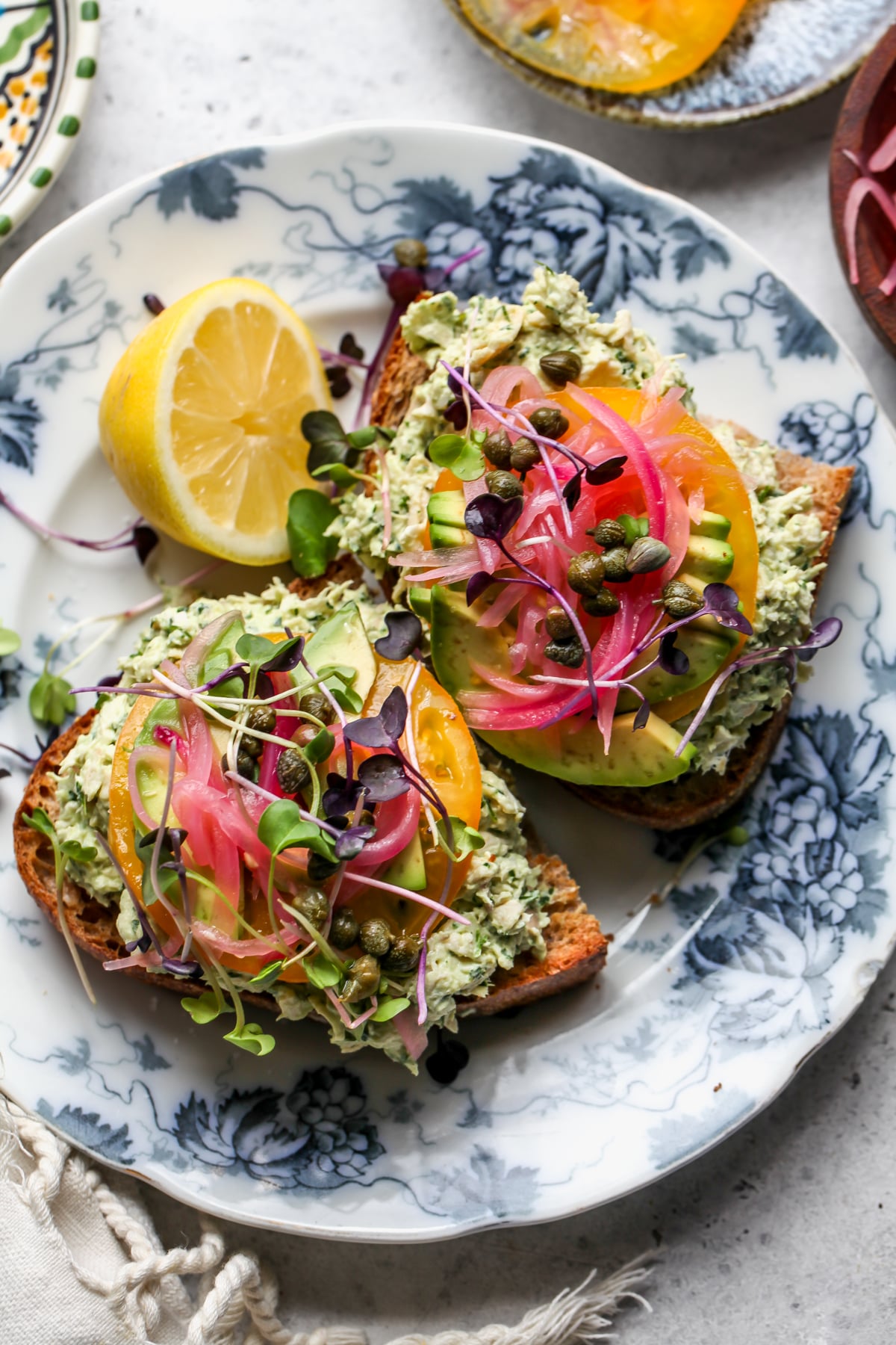 Two slices of bread topped with Green Goddess Chicken Salad, tomato, avocado, pickled red onion, and capers.