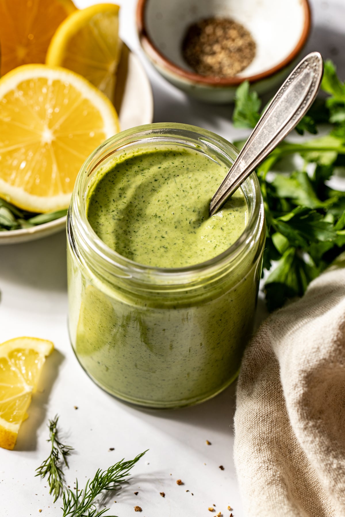 Green goddess tahini dressing in a glass jar with a spoon resting on the right side.
