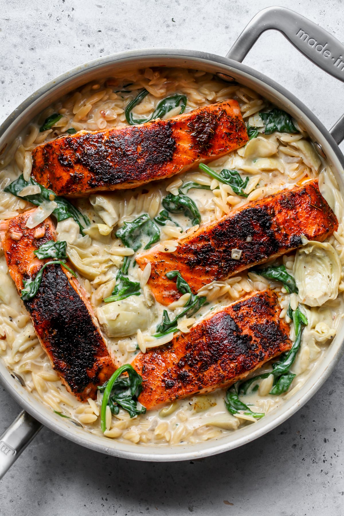 Four cooked salmon fillets nested back into a pan of cooked spinach artichoke orzo.