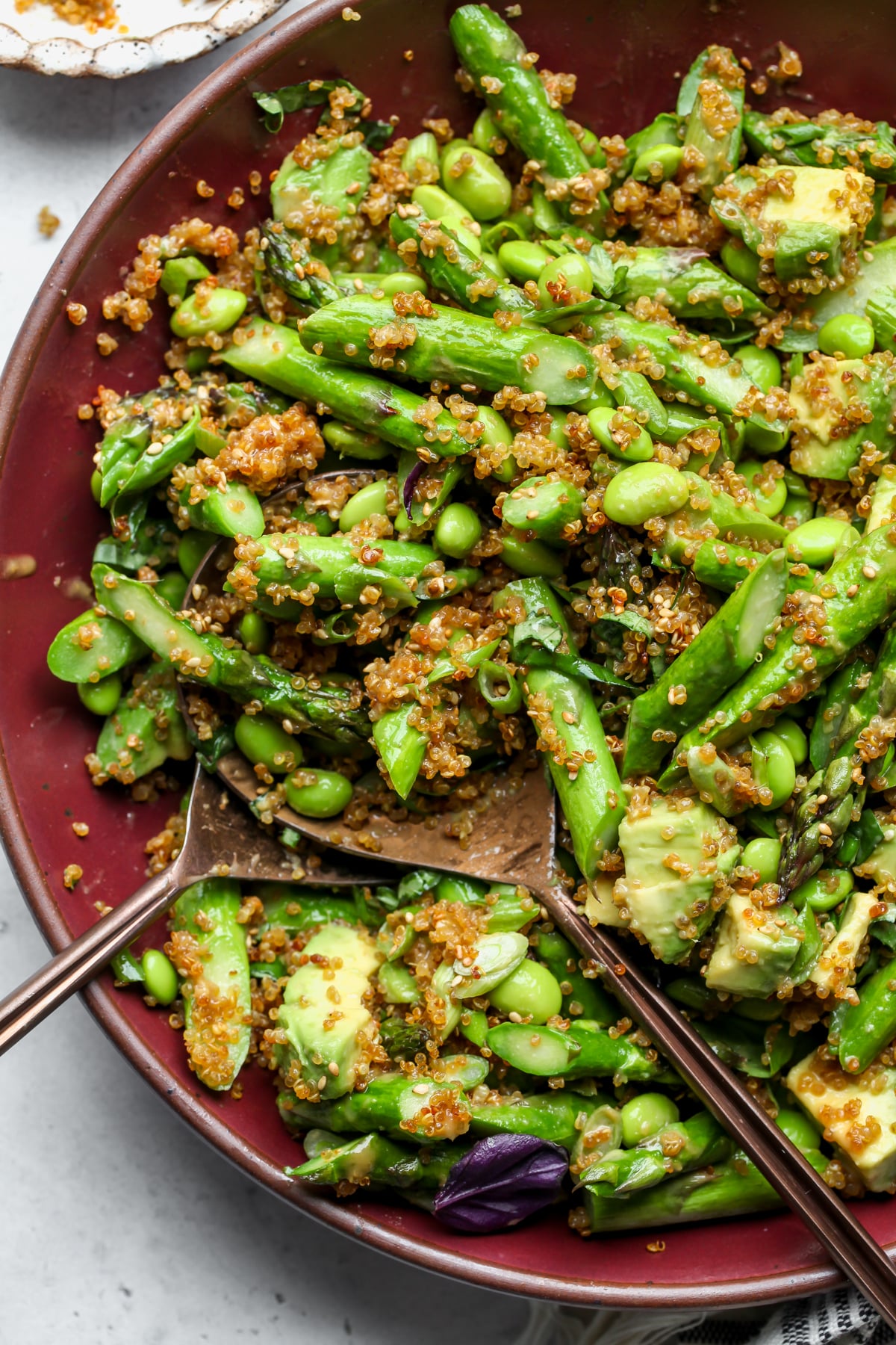 Golden serving spoons tossing an asparagus, quinoa, and edamame salad in a maroon bowl.