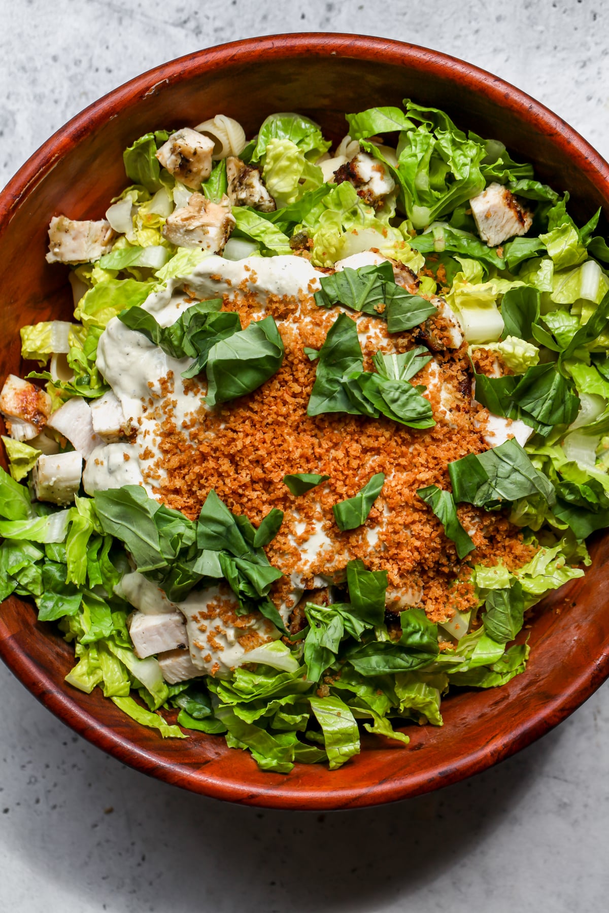Pasta, chicken, romaine, panko, parmesan, and basil leaves being combined in a large wooden bowl.