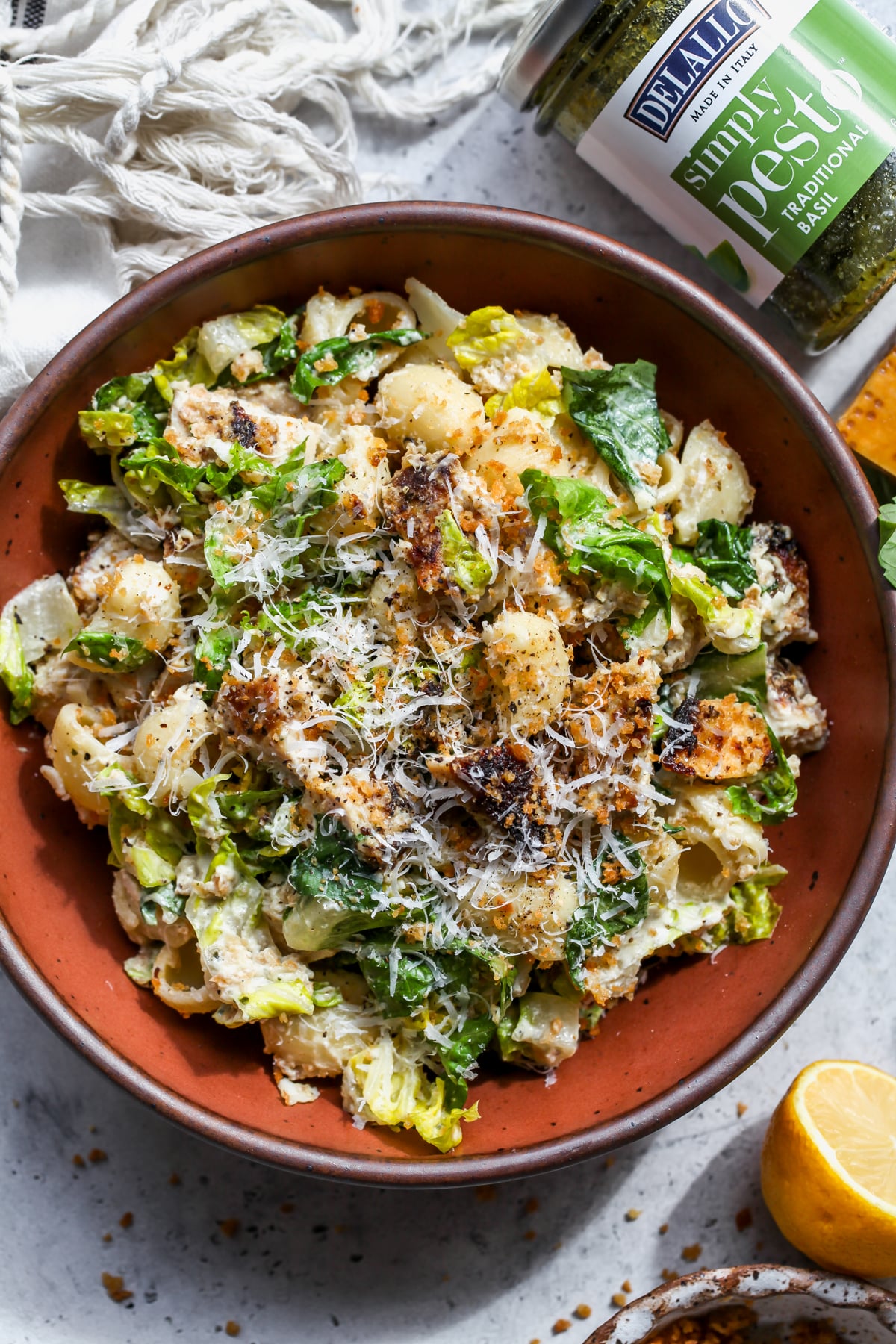 A maroon bowl filled with pesto chicken Caesar salad next to a jar of DeLallo pesto.