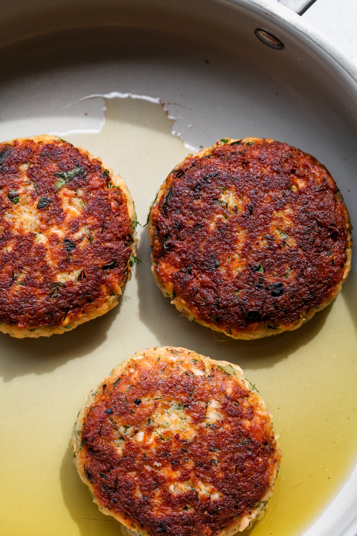 Three salmon patties being seared until golden in a skillet.