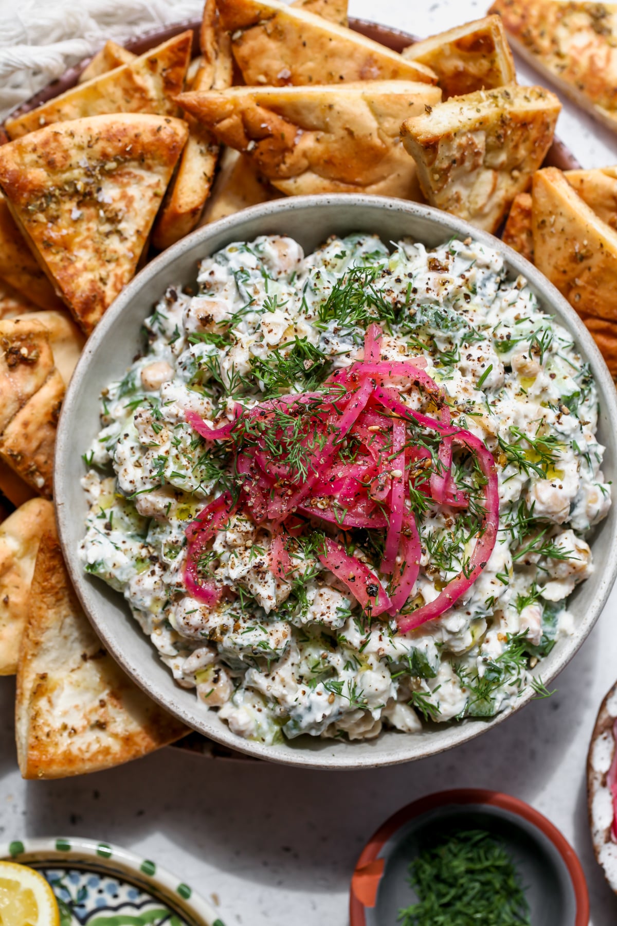Tzatziki chickpea tuna salad topped with pickled red onion in a white bowl surrounded by pita chips.