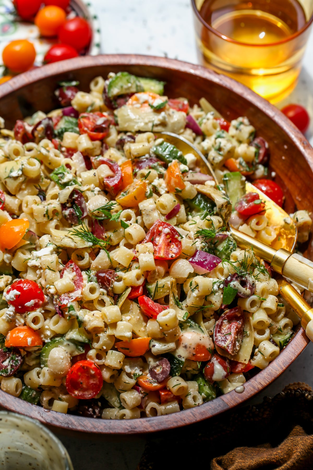 Greek pasta salad being tossed in a wooden serving bowl with two gold serving utensils.