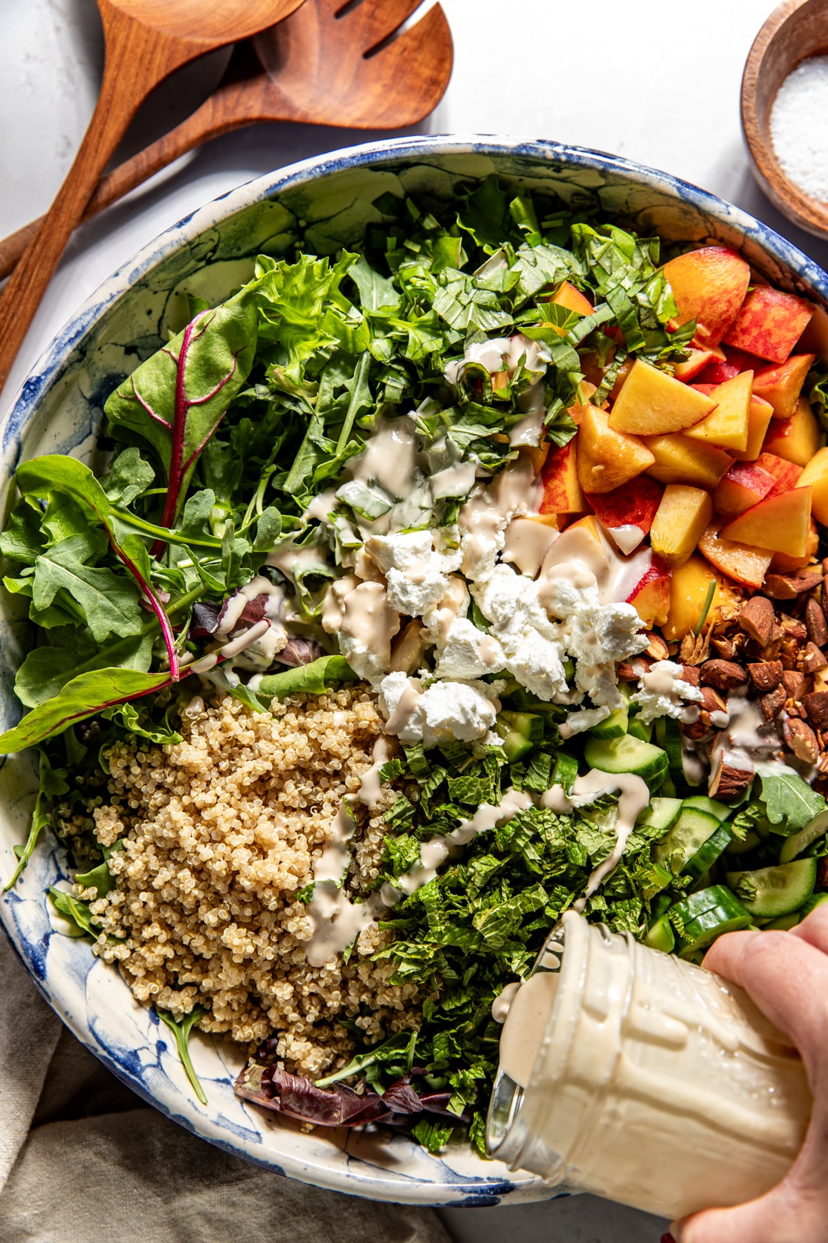 Tahini dressing being poured over peach and goat cheese salad in a large white bowl.