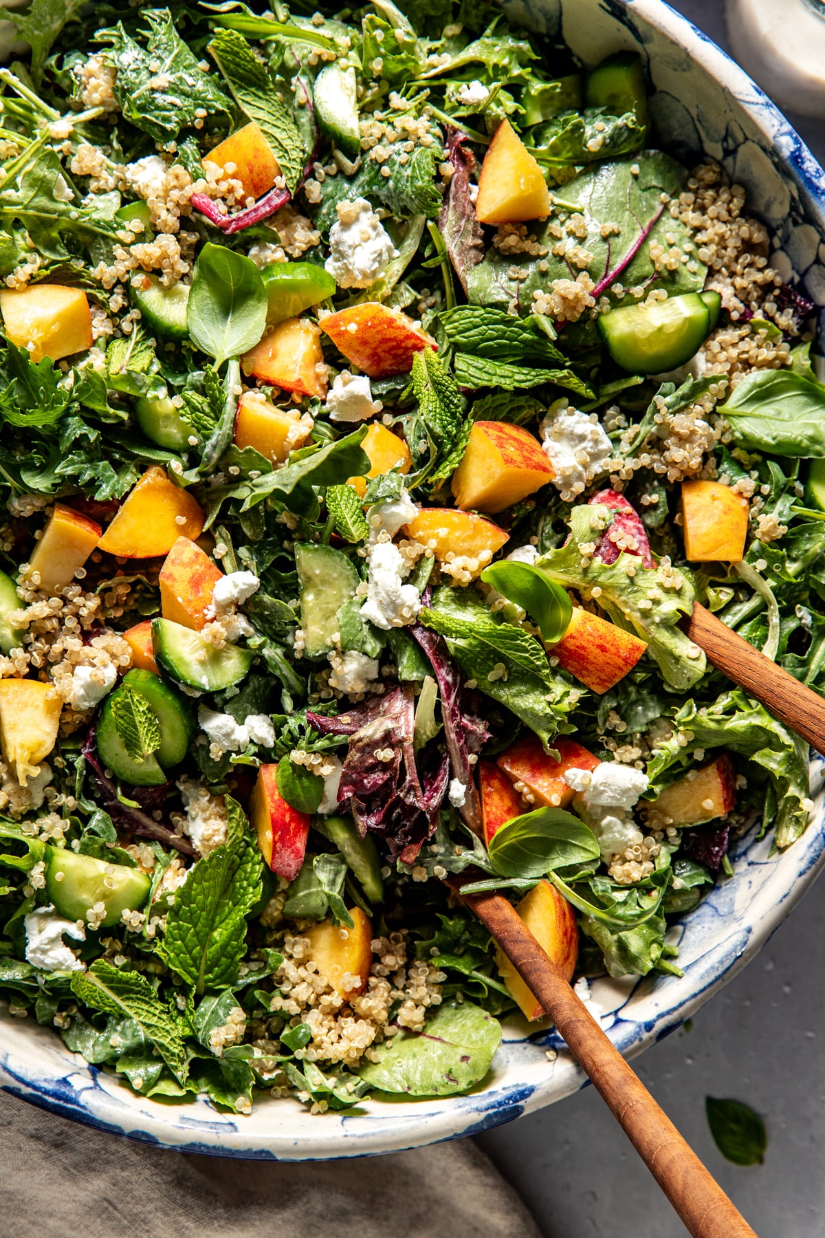 Peach and goat cheese salad being tossed with wooden tongs in a large white speckled bowl.