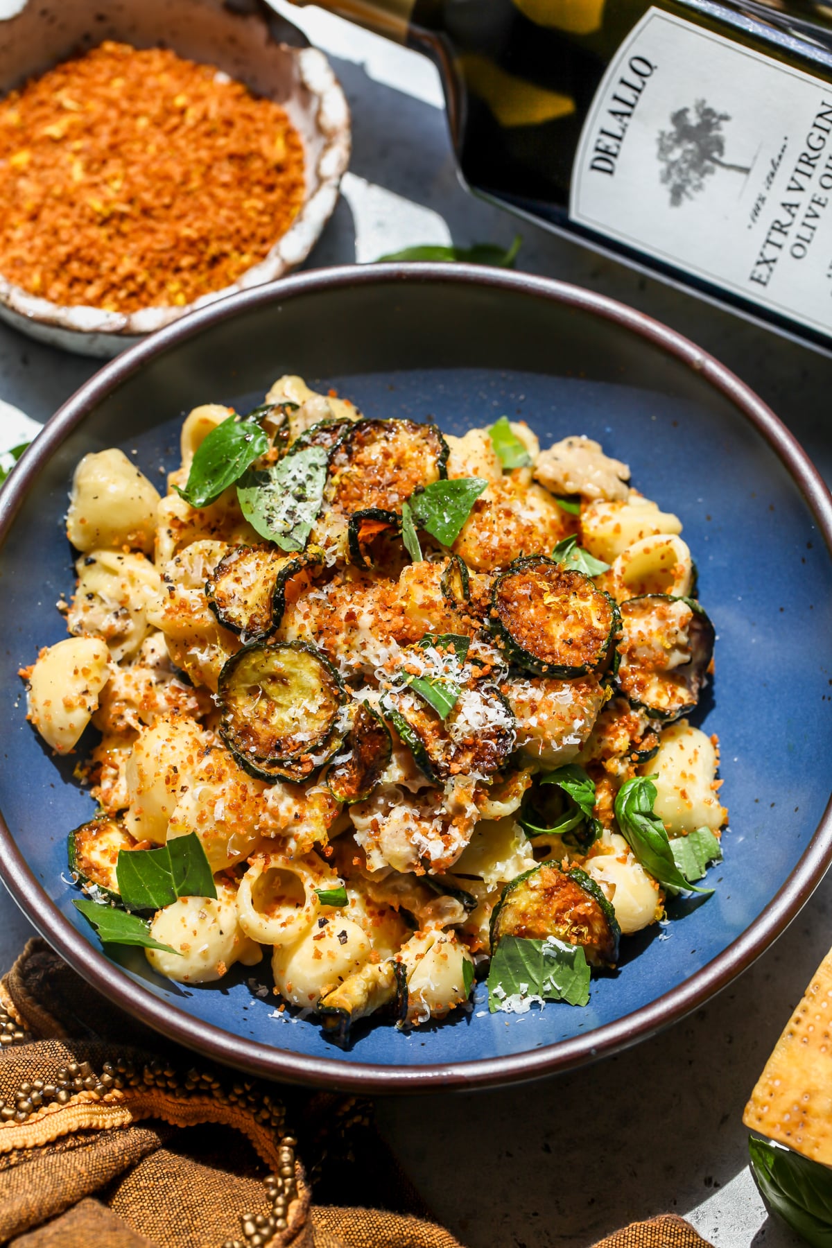 A blue pasta bowl filled with zucchini and sausage pasta topped with parmesan, panko breadcrumbs, and fresh basil. Bottle of extra-virgin olive oil laying next to the bowl.