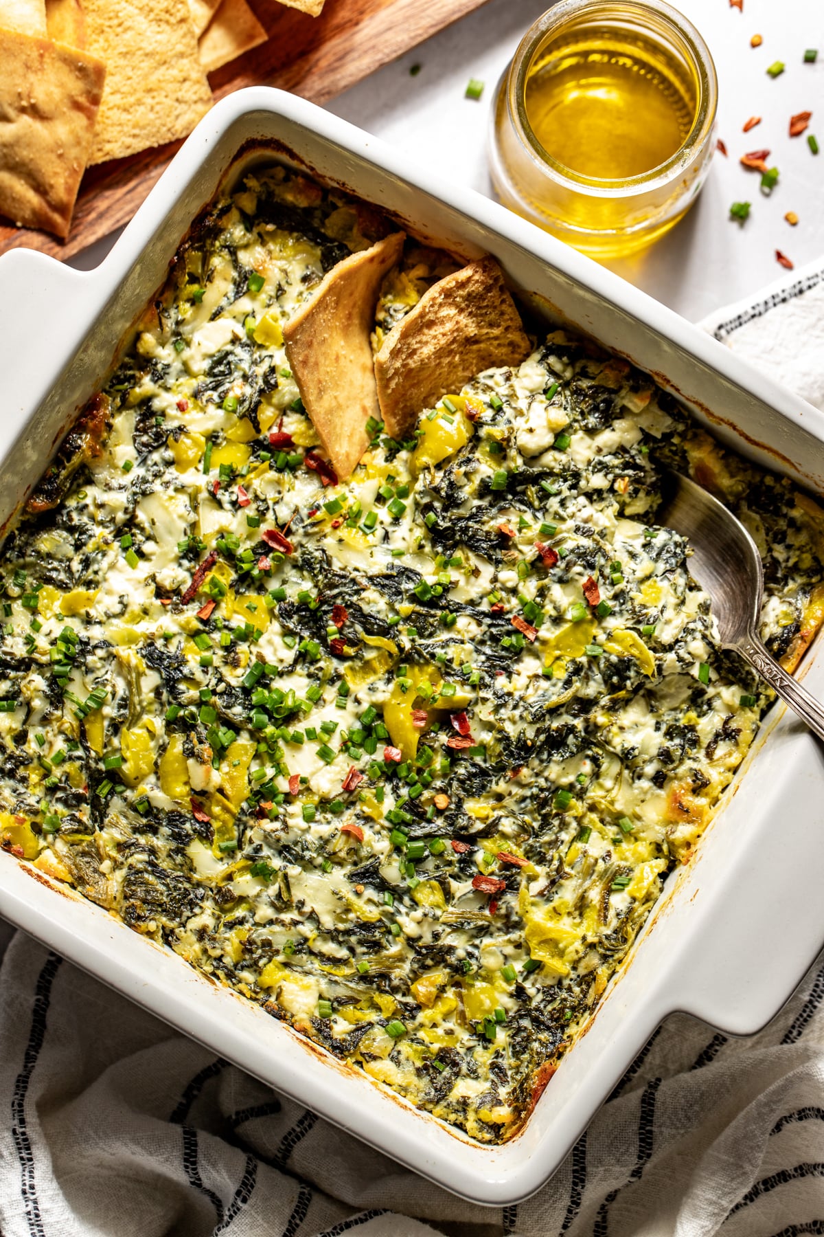 Spinach and pepperoncini baked feta dip shown in a baking dish with pita chips styled in the upper right corner.
