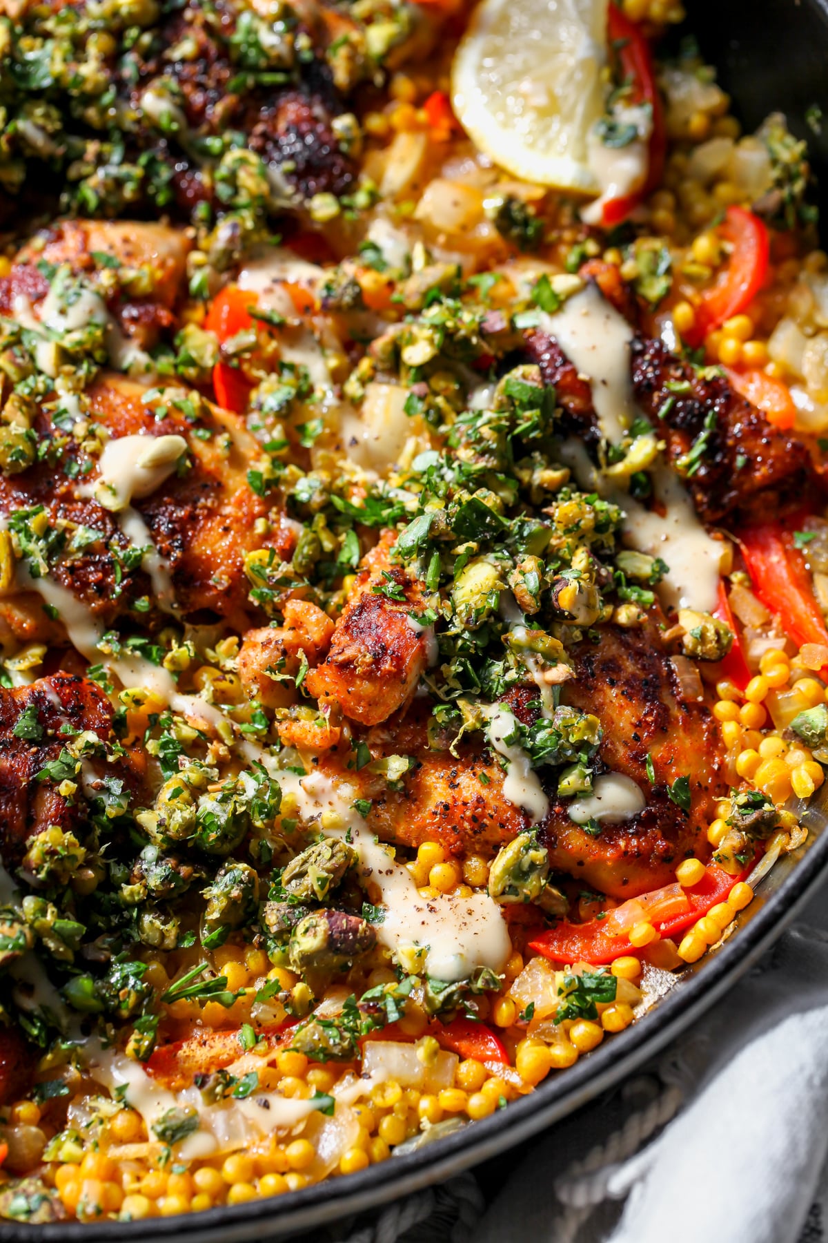 Chicken thigh on a bed of couscous styled in a cast iron skillet topped with pistachios, herbs, and tahini.